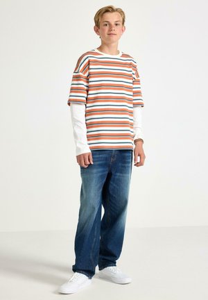Teenage dreng stående, iført orange, hvid og blå stribet kortærmet skjorte over hvide langærmede, blå jeans, hvide sneakers, neutral baggrund.