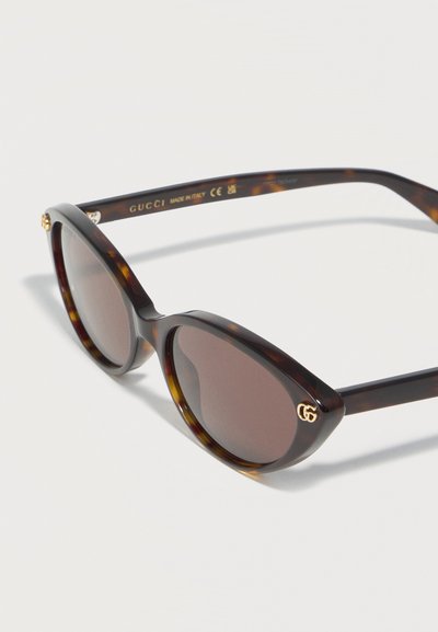 Γυαλιά ηλίου σε σχήμα γάτας με ταρταρούγα, σκοτεινούς φακούς και χρυσό λογότυπο Gucci στην γωνία του δεξιού φακού.