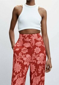 Weißes, geripptes Crop-Top kombiniert mit roten, blütenmusterierten Hosen, die pinke Blumen und grüne Blätter zeigen. Die Hosen haben seitliche Taschen.