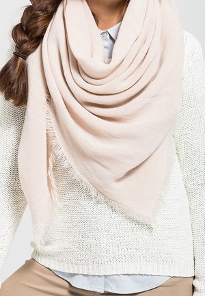 Femme aux cheveux bruns tressés portant un foulard triangulaire beige clair à franges sur un pull en maille blanc et une chemise à col bleu clair.