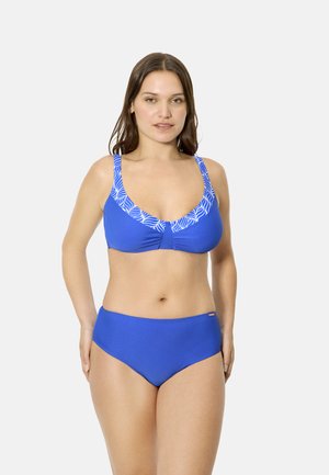 Mujer con un bikini azul con un estampado de hojas blancas en las tiras y el escote de la parte superior, de pie contra un fondo claro y liso.