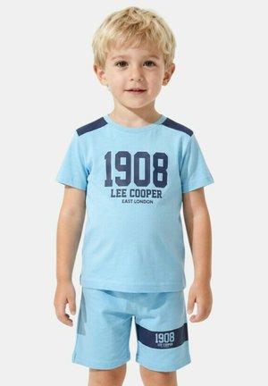 Garçon blond debout, portant un t-shirt bleu clair et un short avec le texte bleu foncé "1908 Lee Cooper East London" et des patchs aux épaules.