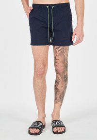 Pantaloni da bagno blu navy con vita elastica e lacci verdi. Presentano un design semplice e una lunghezza sopra il ginocchio. Sandali neri a scivolo.