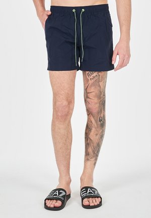 Pantaloni da bagno blu navy con vita elastica e lacci verdi. Presentano un design semplice e una lunghezza sopra il ginocchio. Sandali neri a scivolo.