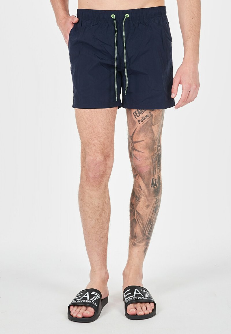 Pantaloni da bagno blu navy con vita elastica e lacci verdi. Presentano un design semplice e una lunghezza sopra il ginocchio. Sandali neri a scivolo.