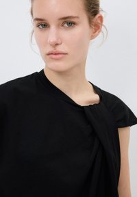 T-shirt nera in morbido cotone con dettaglio annodato sulla spalla e vestibilità rilassata. Tessuto liscio e design minimal.