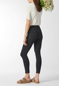 Nainen mustissa kapealahkeisissa farkuissa, valkoisessa lyhythihaisessa paidassa ja ruskeissa kuvioiduissa sandaaleissa, pitäen valkoisten ja keltaisten kukkien kimppua.