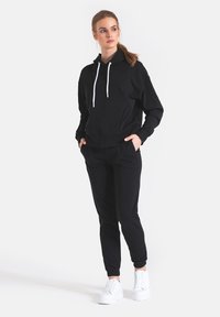 Czarna bluza z kapturem z białymi sznurkami, luźny krój, przednie kieszenie; w komplecie z czarnymi dresami i białymi trampkami, gładka faktura materiału.