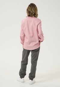 Lyserød langærmet skjorte med en afrundet kant og flæsekrave, parret med grå tapered joggere og hvide sneakers.