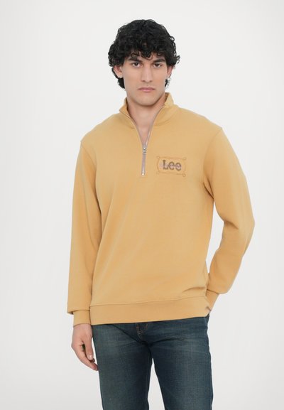 Pull en polaire jaune moutarde avec un col zippé quart, arborant un logo "Leo" brodé en marron sur la poitrine gauche. Texture douce.