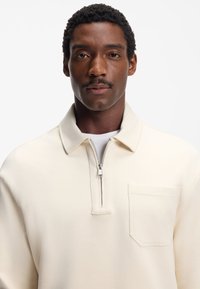 Hellbeiger Zip-Pullover aus weichem Stoff. Verfügt über einen umgelegten Kragen, eine Fronttasche und eine glatte Textur.