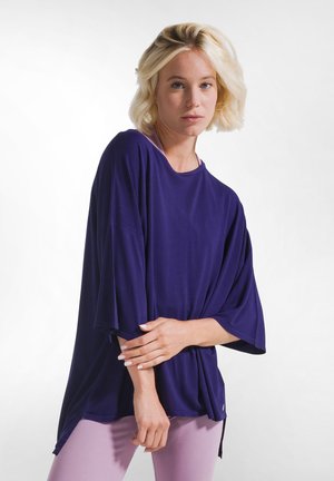 Blondine vrouw met een oversized donkerblauw top en lichtpaarse broek, staand met gekruiste armen tegen een witte achtergrond.