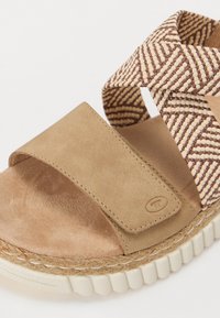Sandale en daim beige avec une lanière à motif tissé marron et crème. Elle dispose d'une semelle texturée et d'arêtes arrondies pour plus de confort.
