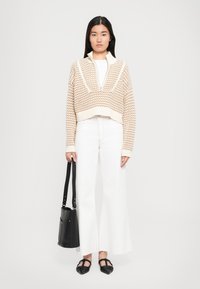 Femme portant un pull zippé en tricot beige, un pantalon large blanc, des ballerines noires pointues et tenant un sac à bandoulière noir devant un fond uni.