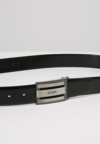 Cinturón de cuero negro con una hebilla de metal cepillado que presenta un diseño de franjas horizontales y el texto grabado "JOOP!". Ajustable con múltiples agujeros.