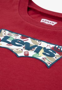 Κόκκινο βαμβακερό T-shirt με τολμηρό μπλε λογότυπο Levi's σε λευκή ετικέτα με πολύχρωμα γραφικά εμπνευσμένα από το φαγητό. Σχεδιασμός με στρογγυλή λαιμόκοψη και απαλό ύφασμα.