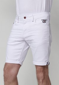 Pantalones cortos de mezclilla blanca con un diseño ajustado. Presenta puños enrollados, acentos de costura roja y un pequeño logo bordado en el bolsillo.