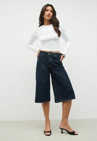 Donkerblauwe denim culottes met een relaxte pasvorm, gecombineerd met een wit aansluitend longsleeve shirt, dat het simpele ontwerp en de casual uitstraling benadrukt.