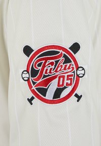 Maillot de baseball blanc avec une texture à rayures. Présente un logo circulaire brodé en rouge, noir et blanc avec des battes croisées et une balle de baseball.