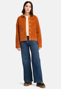 Orange kordjacka med framfickor, stora knappar och en avslappnad passform, ihop med vida blå jeans och kraftiga svarta stövlar.
