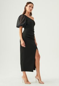 Black one-shoulder dress with a fitted silhouette, featuring a sheer puff sleeve with polka dots and a thigh-high slit. Clear heeled sandals.