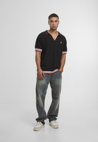 Camisa polo negra de cuello en V con puños a rayas rojas y blancas, combinada con jeans de mezclilla desgastados y zapatillas blancas. Diseño minimalista y corte relajado.