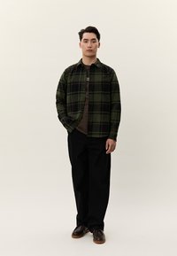 Vyras stovi, dėvėdamas žalią ir juodą languotą marškinių viršų ant rudos spalvos marškinėlių, juodas kelnes ir tamsiai rudas batus, priešais paprastą šviesią foną.