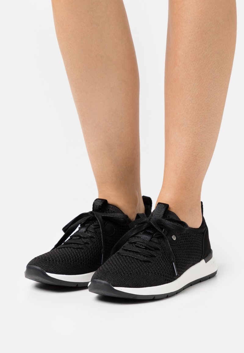 Pieds portant des baskets en tissu tricoté noir avec des semelles blanches, des lacets noués, debout sur une surface blanche uni.