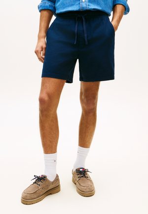 Mężczyzna w granatowych szortach na troczki, białych skarpetkach do kostek, beżowych casualowych butach na sznurowadła oraz z podwiniętą niebieską koszulą dżinsową.