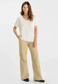 Witte T-shirt met V-hals, gecombineerd met beige broek met wijde pijpen. Gladde stoftextuur met een ontspannen pasvorm. De schoenen zijn zwart. Simpele, casual uitstraling.