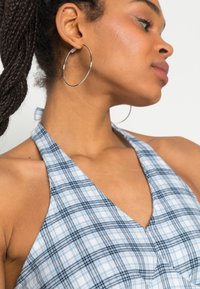 Top de halter em padrão xadrez azul claro, feito de um tecido macio. Apresenta um decote em V profundo e um detalhe de laço nas costas. Brincos grandes de aros visíveis.