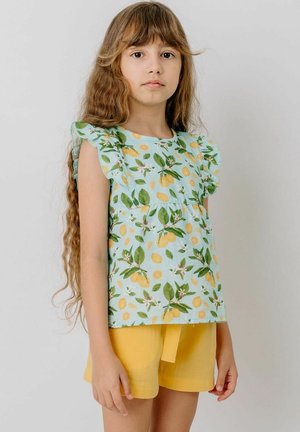 Young girl with long wavy hair wearing a sleeveless blue top with lemon print and yellow shorts, standing against a plain light background.