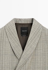 Blazer beige sur mesure avec un fin motif à carreaux noirs en fenêtre et une étiquette noire indiquant Massimo Dutti, fabriqué au Portugal, à l'intérieur du col.