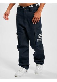 Dark blue cargo pants made of denim, featuring large side pockets, logo accents, and a relaxed fit. Paired with white sneakers.