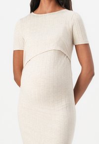 Robe de maternité beige à manches courtes, présentant une texture côtelée et un design croisé au niveau de la poitrine, conçue pour le confort et le soutien.