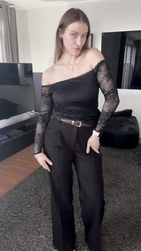 Haut noir à épaules dénudées avec manches en dentelle, associé à un pantalon noir taille haute, ceinturé d'une ceinture marron avec une boucle dorée.