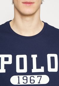 Navy blue cotton t-shirt featuring large white "POLO" text and "1967" in a rounded rectangle below. Classic crew neckline, short sleeves.