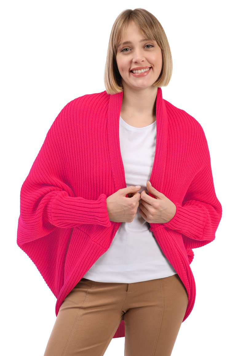 Chal de punto de color rosa brillante con una textura acanalada y mangas anchas, llevado sobre una camisa blanca. El diseño presenta un ajuste holgado y un frente abierto.