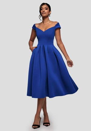 Donna con un vestito midi blu con spalle scoperte e pieghe, in piedi con sicurezza con una mano in tasca e tacchi aperti neri su sfondo grigio.