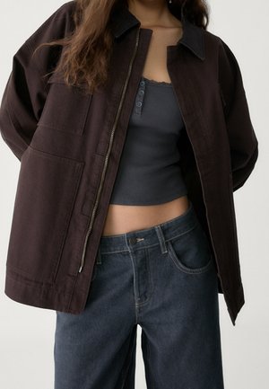 Personne portant une veste zippée marron foncé sur un top court gris foncé et un jean ample taille haute bleu foncé.