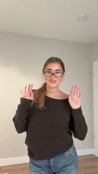 Svart axelbandslös tröja, ljusblå jeans. Person med långt hår, klara glasögon, vinkar med händerna. Enfärgad bakgrund med en dörr.