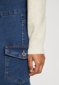 Sötétkék farmeroverall elöl zseb részlettel. Egy krém színű, texturált pulóver ujja részben látható, ribbelt mandzsetta részlettel.