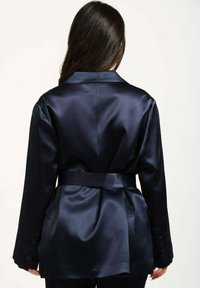 Blazer in satin navy con vita con cintura, maniche lunghe e classico colletto a risvolto. Texture morbida con dettagli a bottoni sulle maniche.