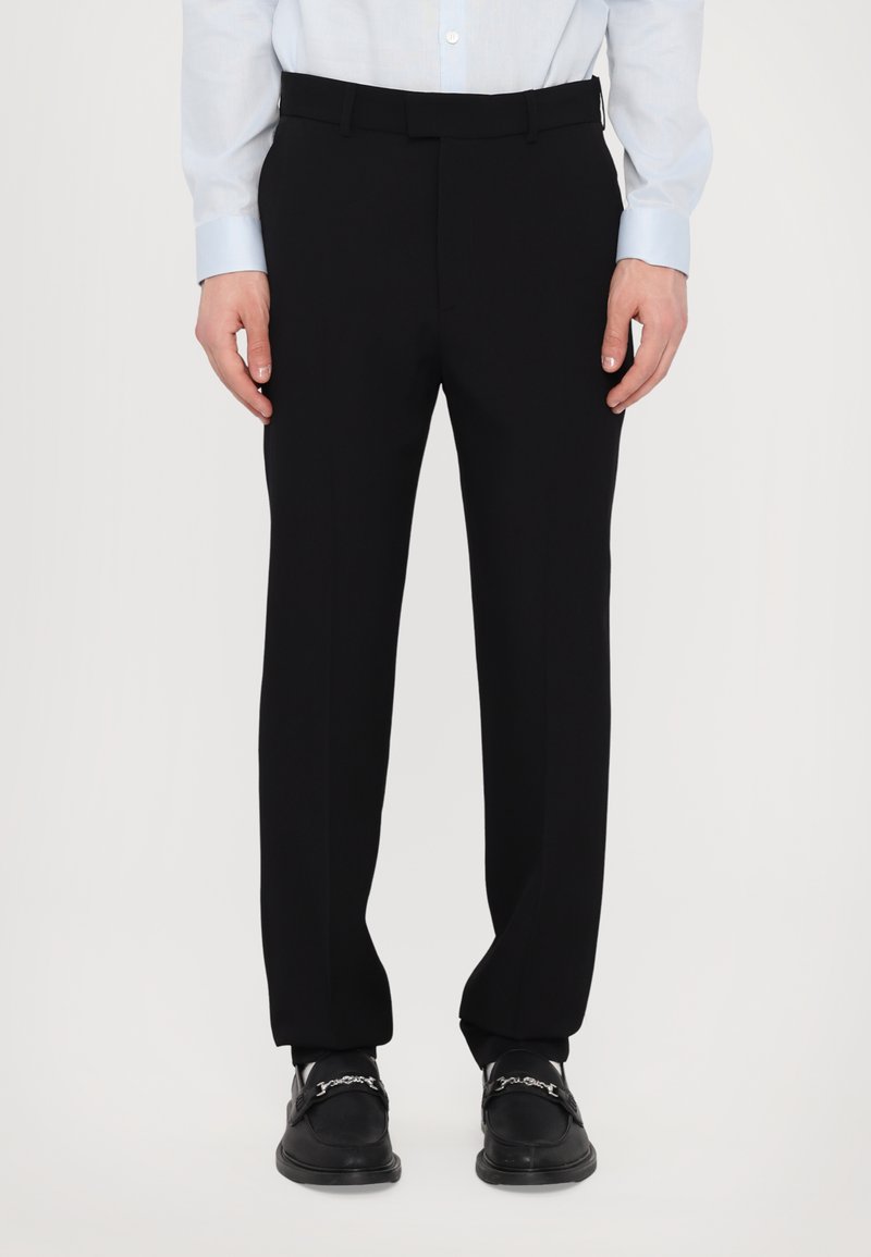 Pantalons noirs ajustés avec une texture lisse, une coupe droite et un design à plat. Portés avec une chemise à manches longues bleu clair.