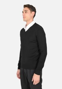 Maglione nero a scollo a V realizzato in un tessuto liscio e morbido, dalla vestibilità aderente, con maniche lunghe, indossato da un modello maschile vestito con una camicia bianca a colletto.