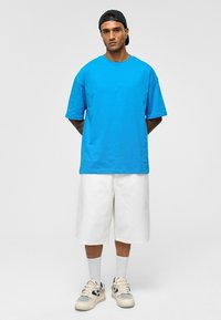 Blauwe katoenen t-shirt met korte mouwen, gecombineerd met witte knielange shorts. Draagt witte sneakers en een zwarte pet.