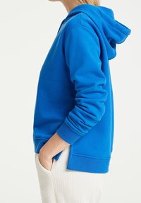 Blue hoodie with a kangaroo pocket, ribbed cuffs, and hem. Soft fabric with a relaxed fit, featured on a model with casual beige pants.