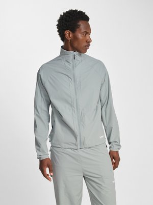 Man draagt een lichtgrijs jack met rits en bijpassende broek, kijkt opzij tegen een effen witte achtergrond.