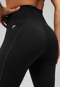Schwarze, hochgeschnitte Leggings mit einer glatten Textur und Kontrastnähten, die ein kleines Logo an der Rückseite in der Nähe des Bundes aufweist.