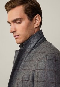 Cappotto di lana grigio con colletto alto, caratterizzato da un motivo a quadri in grigio scuro e borgogna. Sono evidenti la texture liscia e il design strutturato.